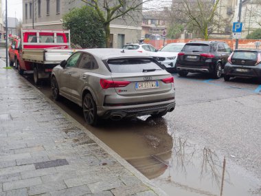 Cremona, İtalya - 3 Şubat 2026 Audi RS Q3 Spor Sırtı, ıslak bir şehir caddesinde tuğla kaldırım kenarına park edilmiş, su birikintisi çıplak ağaçları yansıtıyor ve bulutlu gökyüzü, kentsel kış sürüş atmosferini çağrıştırıyor