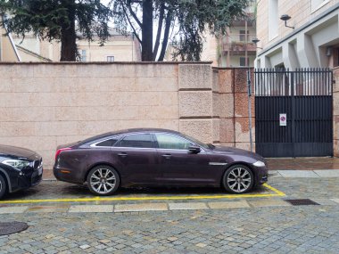 Cremona, İtalya - 3 Şubat 2026 koyu mor bir Jaguar XJ sedan, arka planda büyük metal bir kapısı ve modern mimarisi olan ıslak bir kaldırım taşı caddesinde belirlenmiş bir yere park edilmiştir.