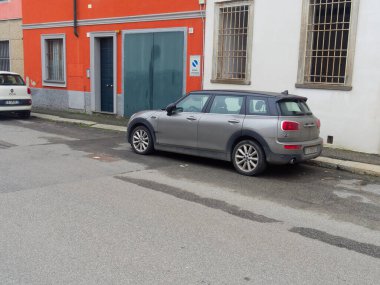 Cremona, İtalya - 3 Şubat 2026 Modern bir gri Mini Cooper Clubman station wagon, turuncu ve beyaz ön cepheli konutların önündeki ıslak asfalt bir caddede park halinde.