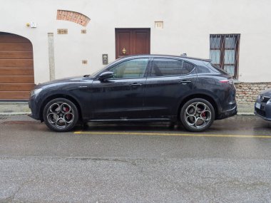 Cremona, İtalya - 3 Şubat 2026 Parlak siyah bir Alfa Romeo Stelvio SUV ıslak asfalt bir yolda, ahşap bir garaj kapısı ve parmaklıklı penceresi olan bir binanın yanında park halinde.