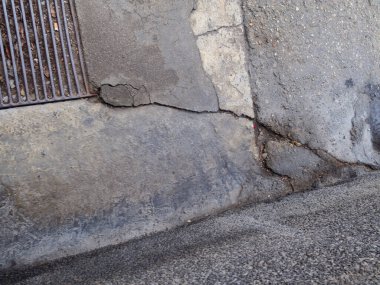 Çürümüş kentsel bozulmayı ve yapısal hasarı ön plana çıkaran, rögarın yanındaki çatlamış beton yüzeyin yakından görüntüsü.