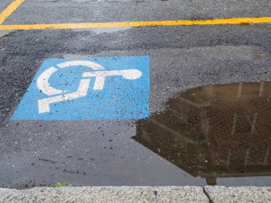 Islak asfalt bir otoparka mavi ve beyaz bir giriş sembolü çizilmiştir. Su birikintisindeki bir binayı yansıtır.