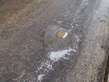 Hasarlı asfalt yol yüzeyinin dokusu gökyüzünü ve çevreyi yansıtan birkaç su birikintisi ve çatlakla dolu.