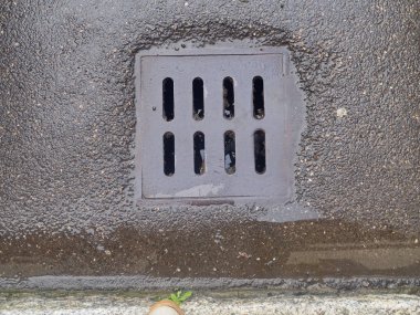 Koyu, ıslak asfalt bir yüzeye yakın çekim. Üzerinde görünür su birikintisi olan dikdörtgen metal bir drenaj kapağı var.