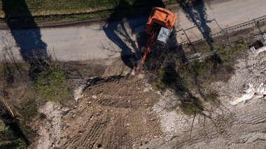 Bir turuncu kazıcı etkin bir şekilde kirli bir inşaat alanında çalışıyor, kovasıyla toprağı ve molozları hareket ettiriyor.