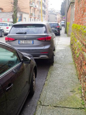 Cremona, İtalya - 26 Şubat 2026 Gri bir MG SUV, arka planda diğer araçların da görüldüğü, çatlamış beton kaldırım ve yosun kaplı tuğla duvarın yanındaki dar bir caddede park halinde.