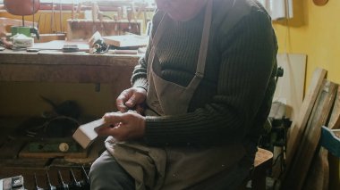 Cremona, İtalya 'da yaşlı bir zanaatkar, ahşap bir parçayı dikkatlice şekillendirir, muhtemelen bir keman parşömeni için, aletler ve malzemelerle çevrili.