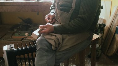 Tozlu bir atölyedeki bir zanaatkar dikkatle bir odun parçasını şekillendirir, muhtemelen bir keman parşömeni için, odaklanmış hassasiyetle.