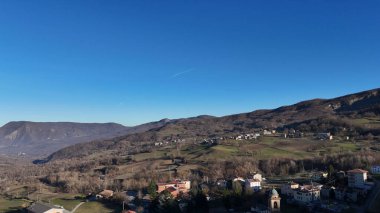 Açık mavi bir gökyüzü, evlerle ve yemyeşil çayırlarla benekli İtalyan kırsalının üzerine uzanır.
