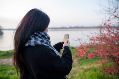 Elinde akıllı telefon tutan bir kadın, kırmızı çiçekli bir nehrin yakınındaki çevreyi belgeliyor, manzaralı bir rıhtımda doğa ile keyif ve bağlantı buluyor.
