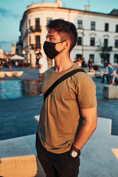 Young man standing in the city center, looking away, wearing the face mask to avoid virus infection and to prevent the spread of disease in time of coronavirus