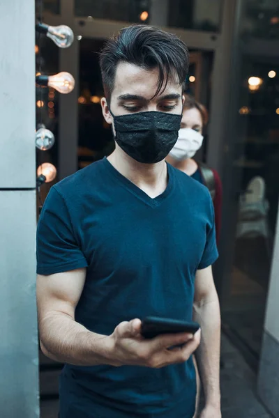 Young man standing in front of restaurant using smartphone wearing the face mask to avoid virus infection and to prevent the spread of disease in time of coronavirus