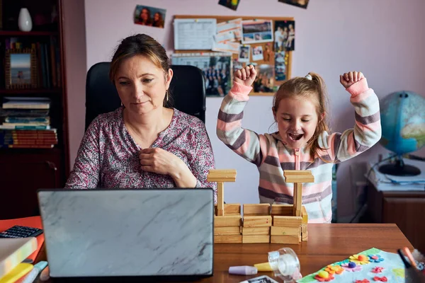 Woman mother working doing her job remotely during video chat call stream online course webinar on laptop from home while her daughter playing with bricks toy. Woman sitting at desk in front of computer looking at screen