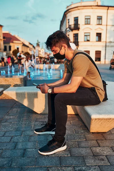 Молодой Человек Сидящий Центре Города Вечером Используя Смартфон Носит Маску — стоковое фото