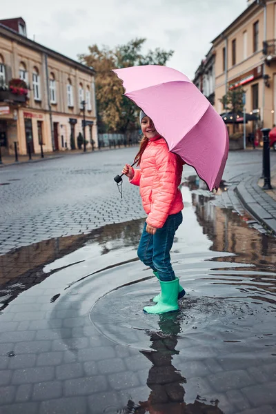 Child holding big pink umbrella walking through the puddle in a downtown on rainy gloomy autumn day