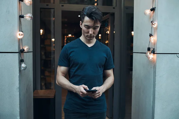 Young man standing in front of restaurant in the city center holding smartphone looking at screen