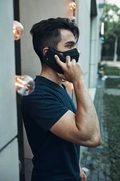 Young man standing in front of restaurant talking on phone using smartphone wearing the face mask to avoid virus infection and to prevent the spread of disease in time of coronavirus