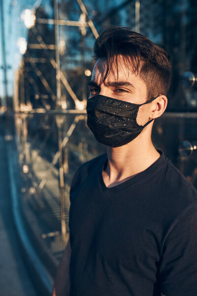 Young man standing in the city center at glass store front in the evening looking away wearing the face mask to avoid virus infection and to prevent the spread of disease in time of coronavirus