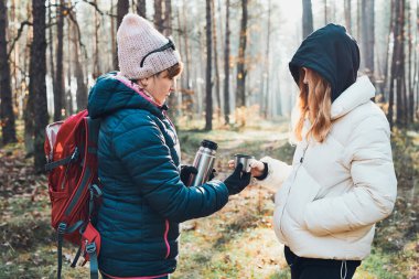 Sırt çantalı anne ve kızı sonbahar tatilinde soğuk sonbahar gününde termostan sıcak bir içecek dolduruyorlar. Aktif kadınlar ormanda aktif olarak vakit geçiriyorlar.