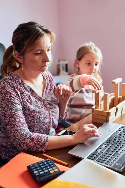 Woman mother working doing her job remotely during video chat call stream online course webinar on laptop from home while her daughter playing with bricks toy. Woman sitting at desk in front of computer looking at screen