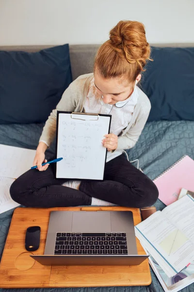 Young woman student having classes, learning online, watching lesson remotely, listening to professor, talking with classmates on video call from home during quarantine. Young girl using laptop, headphones, books, manuals sitting on bed