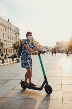 Şehir merkezinde elektrikli scooter kullanan genç bir kadın. Virüs enfeksiyonunu önlemek ve koronavirüs zamanında hastalığın yayılmasını önlemek için yüz maskesi takan kadın. Samimi insanlar, gerçek anlar, otantik durumlar.