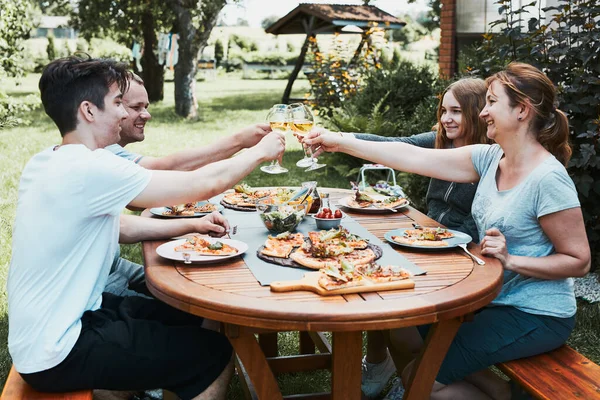 Friends making toast during summer picnic outdoor dinner in a home garden. Close up of people holding wine glasses with white wine over table with pizza, salads and fruits. Dinner in a orchard in a backyard