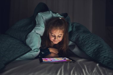 Küçük kız tablet bilgisayarda izleyip, oynayıp, hikayeler dinleyerek eğleniyor. Çocuk yatmadan önce yorganın altında tablet kullanarak eğleniyor.