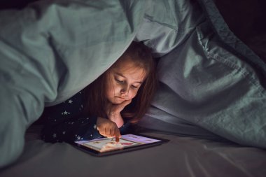 Küçük kız tablet bilgisayarda izleyip, oynayıp, hikayeler dinleyerek eğleniyor. Çocuk yatmadan önce yorganın altında tablet kullanarak eğleniyor.