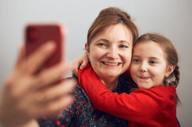Anne ve küçük kızı cep telefonuyla video görüşmesi yapıyor. Mesafeyi koruyor. Kadın ve küçük kız akrabalarıyla konuşuyor. Neşeli aile akıllı telefon kullanarak selfie çekerken eğleniyor. Aileye uzaktan bağlanma