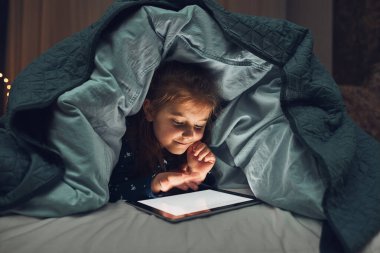 Küçük kız tablet bilgisayarda izleyip, oynayıp, hikayeler dinleyerek eğleniyor. Çocuk yatmadan önce yorganın altında tablet kullanarak eğleniyor.