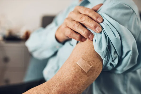 Senior man patient holding shirt sleeve up with a plaster in place of injection of vaccine. Covid-19 or coronavirus vaccination. Man wearing face mask