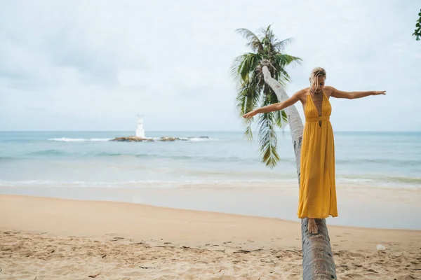 ビーチでヤシの木の幹に沿って歩いて黄色のドレスの幸せな若い女性 — ストック写真