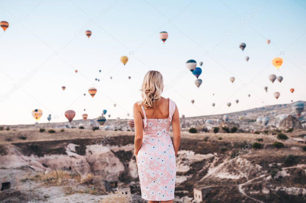 Young woman in dress and hat on a mountain top enjoying wonderful view of the sunrise and balloons in Cappadocia. Travel in Turkey concept