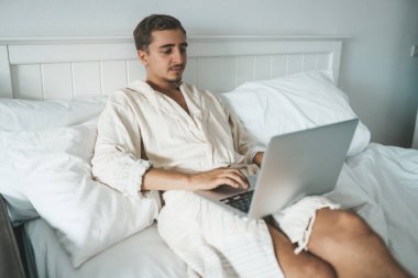 Adam yatakta yatıp dizüstü bilgisayarla çalışıyor.