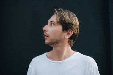 Beyaz tişört giyen bir adamın yan görünüm portresi yukarı bakıp siyah duvara şaşırmış.