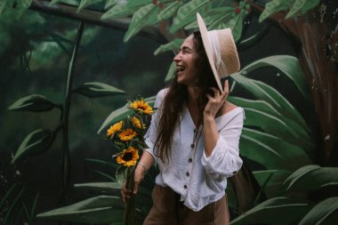 Şapkalı mutlu kadın elinde çiçekle yeşil duvarda duruyor.