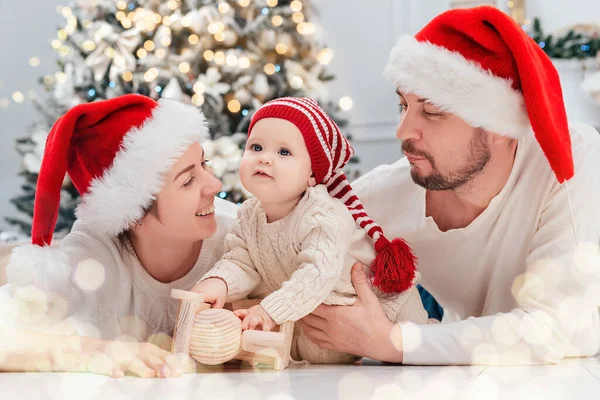 Happy family under Christmas tree. baby boy in Santa Claus hat with gifts under Christmas tree with many gift boxes presents. Happy Holidays, New year. Cozy warm winter evening at home. Xmas time