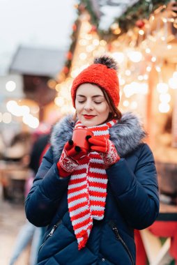 Noel pazarında punç içen genç bir kadın. Caddede dikilmiş sıcak şarap, çay ya da kakao içen, kışın açık havada sıcak kahve içen güzel, kırmızı şapkalı bir kız. Noel ışıkları