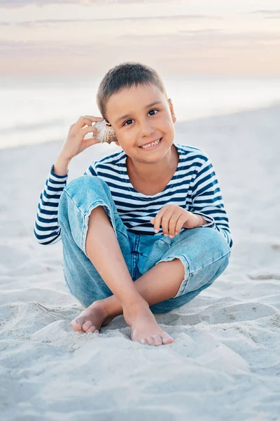 可愛い男の子が浜辺で貝殻の海の音を聞いている 海辺の縞模様のTシャツの子供 夏休みだ 幸せな子供は日没時にビーチで遊んでいます 幸せな子供時代 — ストック写真