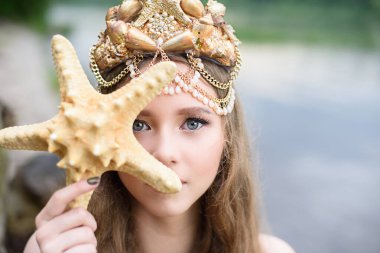 Fantezi kadın gerçek deniz kızı deniz tanrıçası altın kuyruklu deniz tanrıçası gün batımında kayalarda oturuyor altın saçlı taç inci kabuk takıyor. Deniz kızı kıyıda oturuyor. fantezi kavramı.