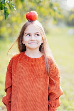 Dışarıda organik kırmızı elma yiyen bir kızın portresi. Hasat Konsepti. Sonbaharda çiftlikte elma toplayan bir çocuk. Çocuk ve Ekoloji. Sağlıklı beslenme bahçesi yemekleri. Kafasında elma tutan kız.