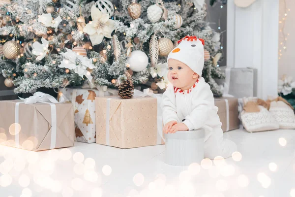 Happy Holidays, New year. Little child under Christmas tree. baby boy in snowman hat with gifts under Christmas tree with many gift boxes presents. Cozy warm winter evening at home. Xmas time