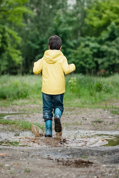 水溜りに飛び込むぬれた子供 路上で楽しい 夏は気温が上がる スプラッシュ 水の滴 防水ブーツは雨の中で水たまりや泥の中にジャンプします 幸せな子供時代 — ストック写真