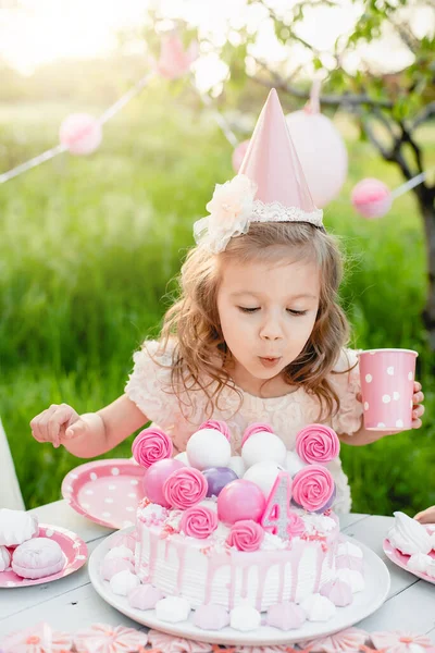 Happy birthday little girl making wish blowing candles on cake with pink decor in beautiful garden. 4 years birthday. child eat happy birthday pink cupcake. colorful pastel decoration outdoor