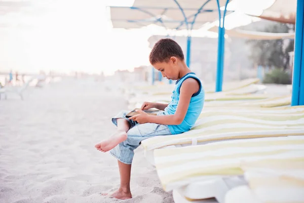青いTシャツを着た少年がビーチで一人で海の近くのベンチに座り タブレットで遊んでいます 幸せな家族旅行 子供と夏休み — ストック写真