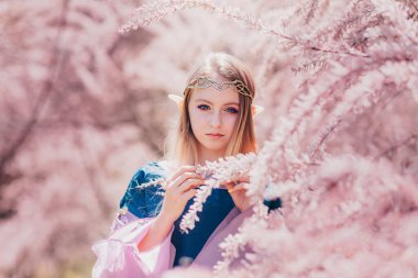 Ormanda poz veren mavi gözlü güzel, romantik sarışın kız. Peri masalı kızı. Cosplay karakteri. Uzun kulaklı elf kızı portresi pembe çiçeklere dokunur. Peri masalı ve fantezi işi.