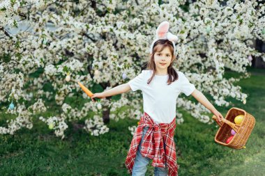 Paskalya yumurtası avı. Bahçede yumurta toplamak için koşan tavşan kulaklı bir kız çocuğu. Paskalya geleneği. Renkli yumurtalarla dolu sepeti olan bebek.