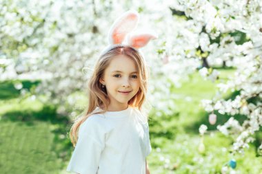 Paskalya yumurtası avı. Bahçede yumurta toplamak için koşan tavşan kulaklı bir kız çocuğu. Paskalya geleneği. Renkli yumurtalarla dolu sepeti olan bebek.