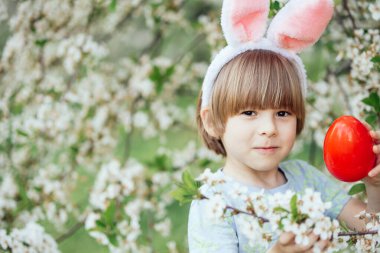 Paskalya yumurtası avı. Bahçede yumurta toplamak için koşan tavşan kulaklı çocuk. Paskalya geleneği. Renkli yumurtalarla dolu sepeti olan bebek.